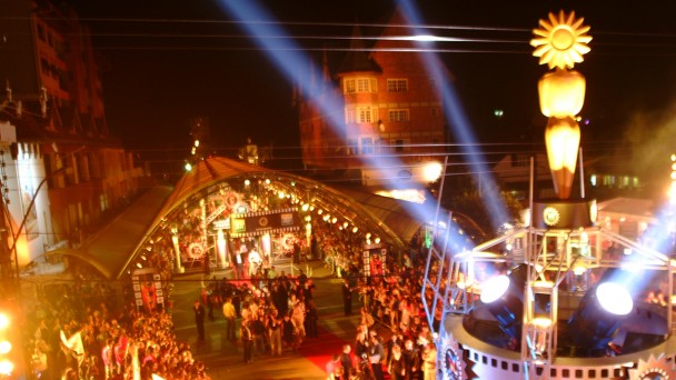 Vista de cima da entrada do festival de cinema de Gramado, com público aglomerado em frente ao tapete vermelho. Um grande Kikito iluminado aparece em destaque no primeiro plano