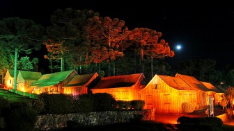 Casas típicas de madeira da serra gaúcha iluminadas à noite, sob as araucárias, em noite de lua cheia.
