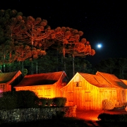 Casas típicas de madeira da serra gaúcha iluminadas à noite, sob as araucárias, em noite de lua cheia.